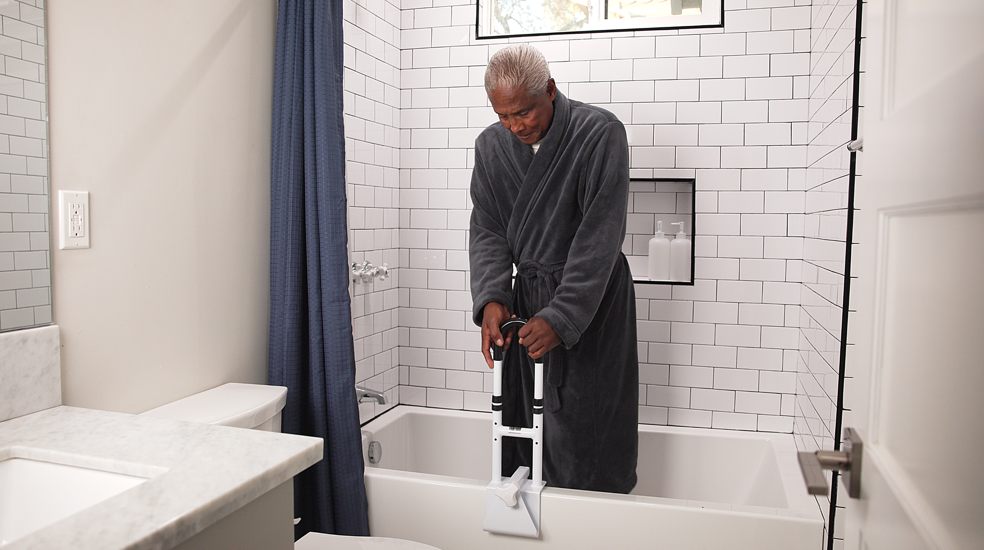 Man in a gray robe using a handheld showerhead in a bathroom.