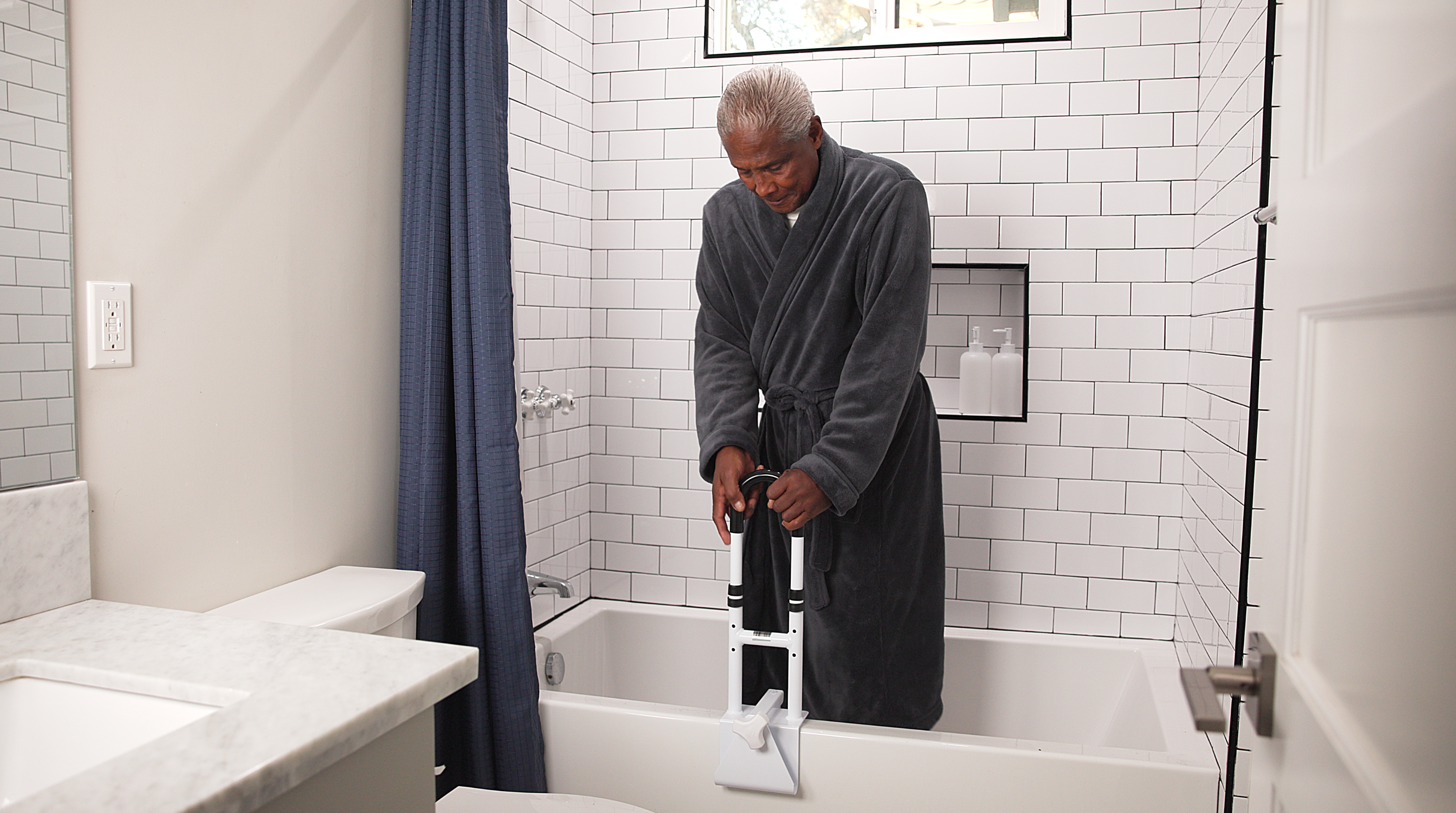 Man in a gray robe using a handheld showerhead in a bathroom.
