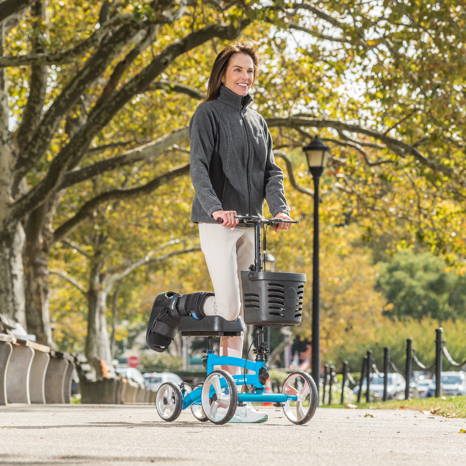 Woman riding a knee scooter in a park with trees and a lamp post in the background.
