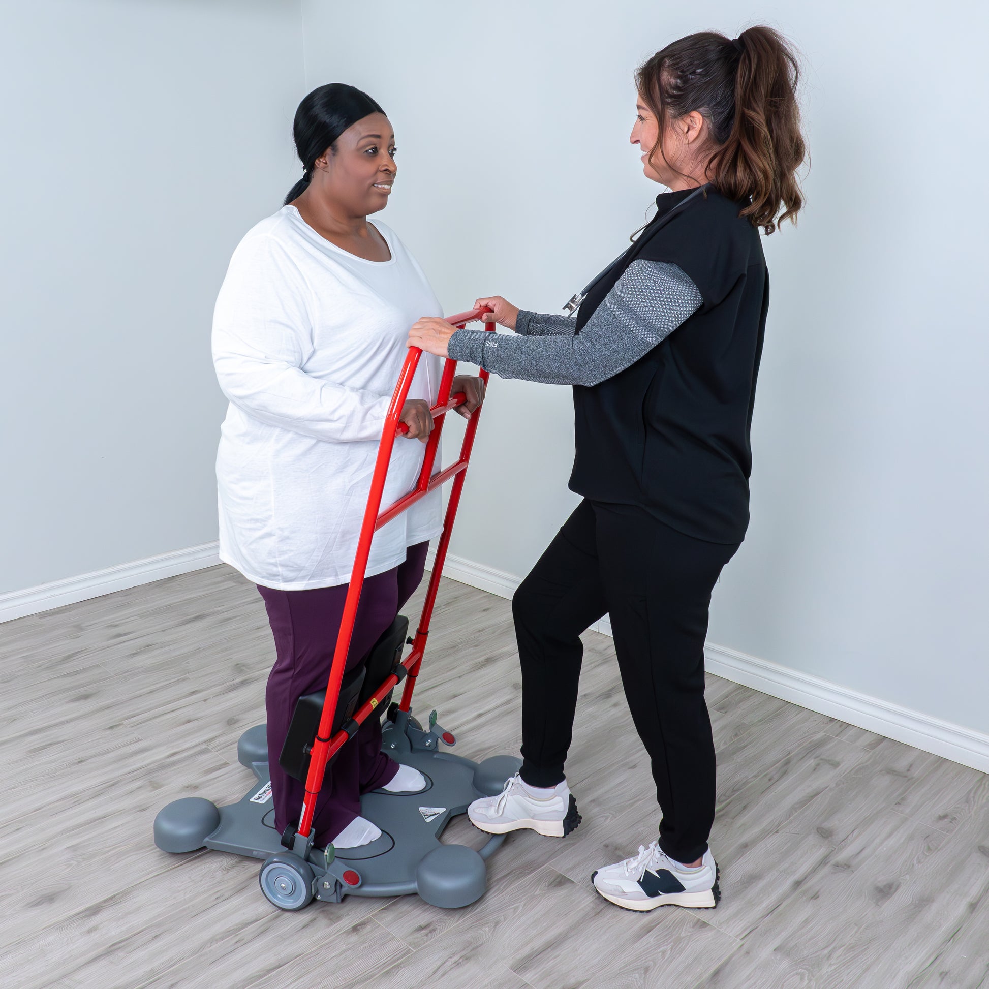 Two women using a mobility aid on a wooden floor with a white wall background